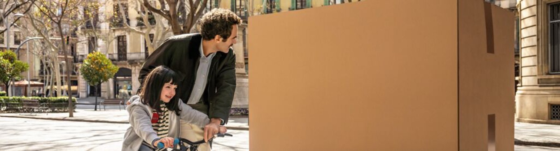 A dad looks at a big parcel while he was teaching his daughter how to ride a bike