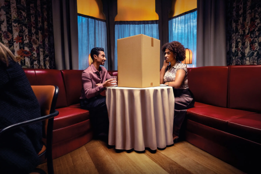 Two people dining in a restaurant, separated by a large cardboard box on the table—symbolizing a broken customer experience. A visual metaphor used by GLS to highlight the importance of smooth, personalized delivery, a key topic at Tech For Retail 2025.