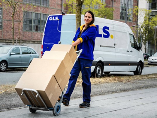 Livreuse GLS transportant plusieurs colis sur un diable, avec un fourgon de livraison GLS en arrière-plan.