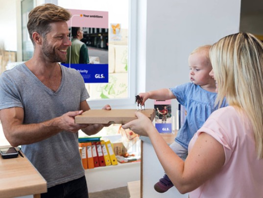 Une femme avec un bébé récupère un colis auprès d’un commerçant dans un Point Relais GLS.